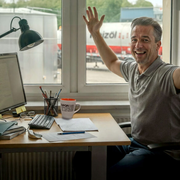 Mann sitzt jubelnd an einem Schreibtisch im Büro. Vor ihm Monitor und Lampe, im Hintergrund LKWs vor dem Fenster. {{brizy_dc_image_alt imageSrc=